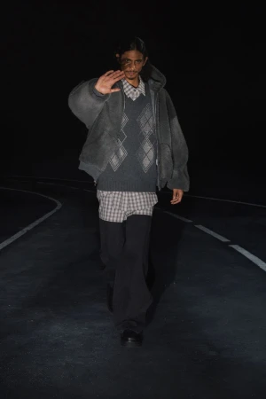 Adult male poses on dark road wearing a grey hooded jacket, patterned shirt, sweater vest, and black pants in a fashion editorial.