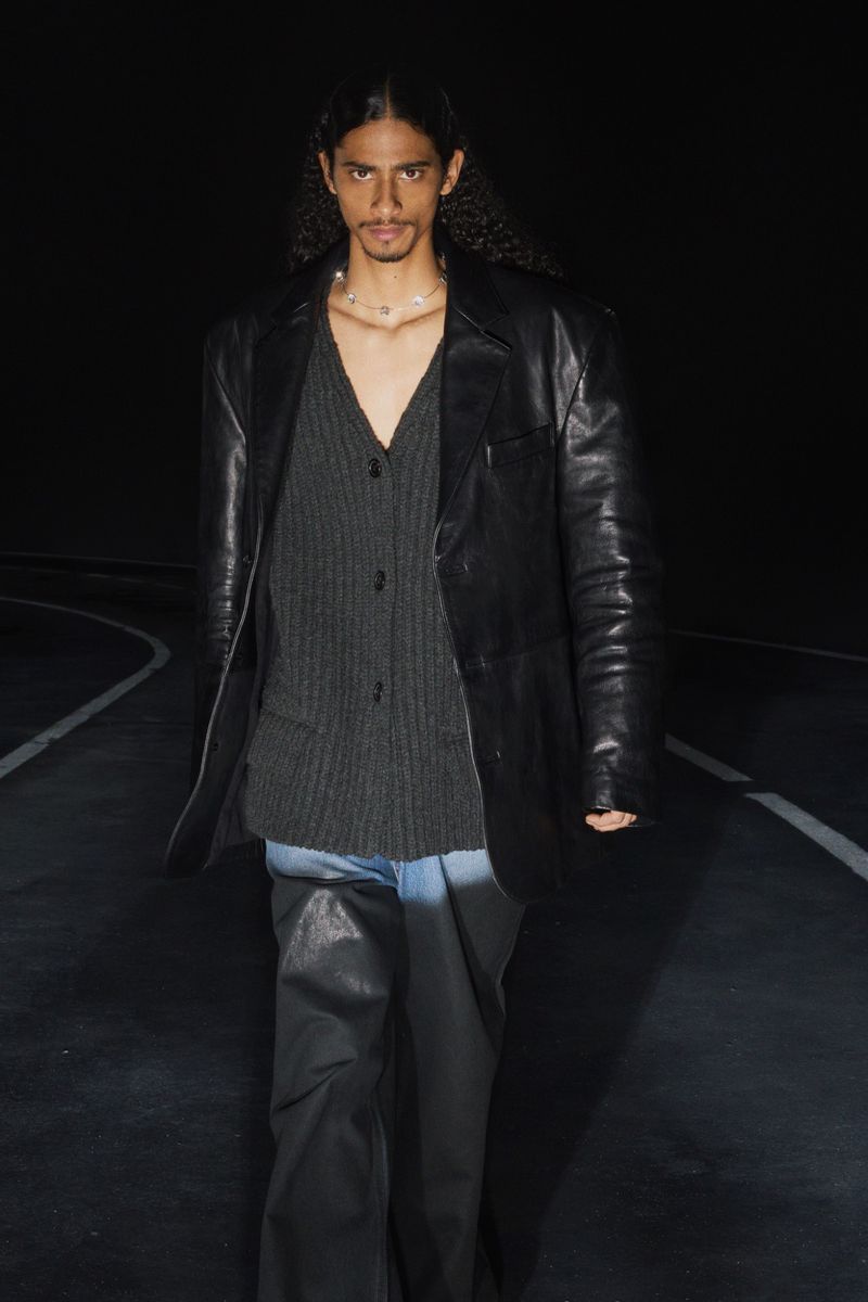 Man in black leather jacket and gray cardigan walking on dark surface with white lines in a fashion editorial shot, moody lighting.