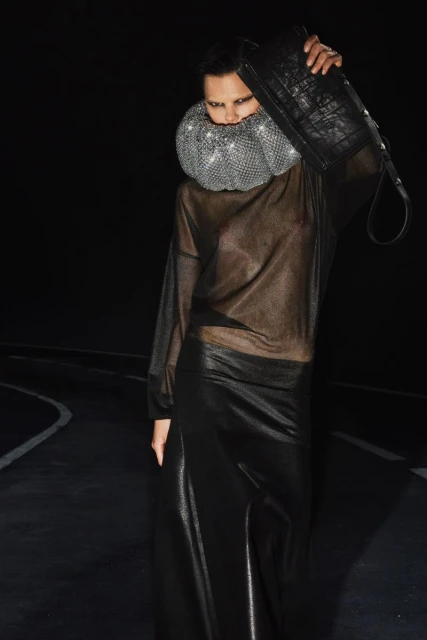 A woman in a sheer top and a long skirt, accessorized with a beaded collar and a black bag, stands against a dark backdrop with a floor.