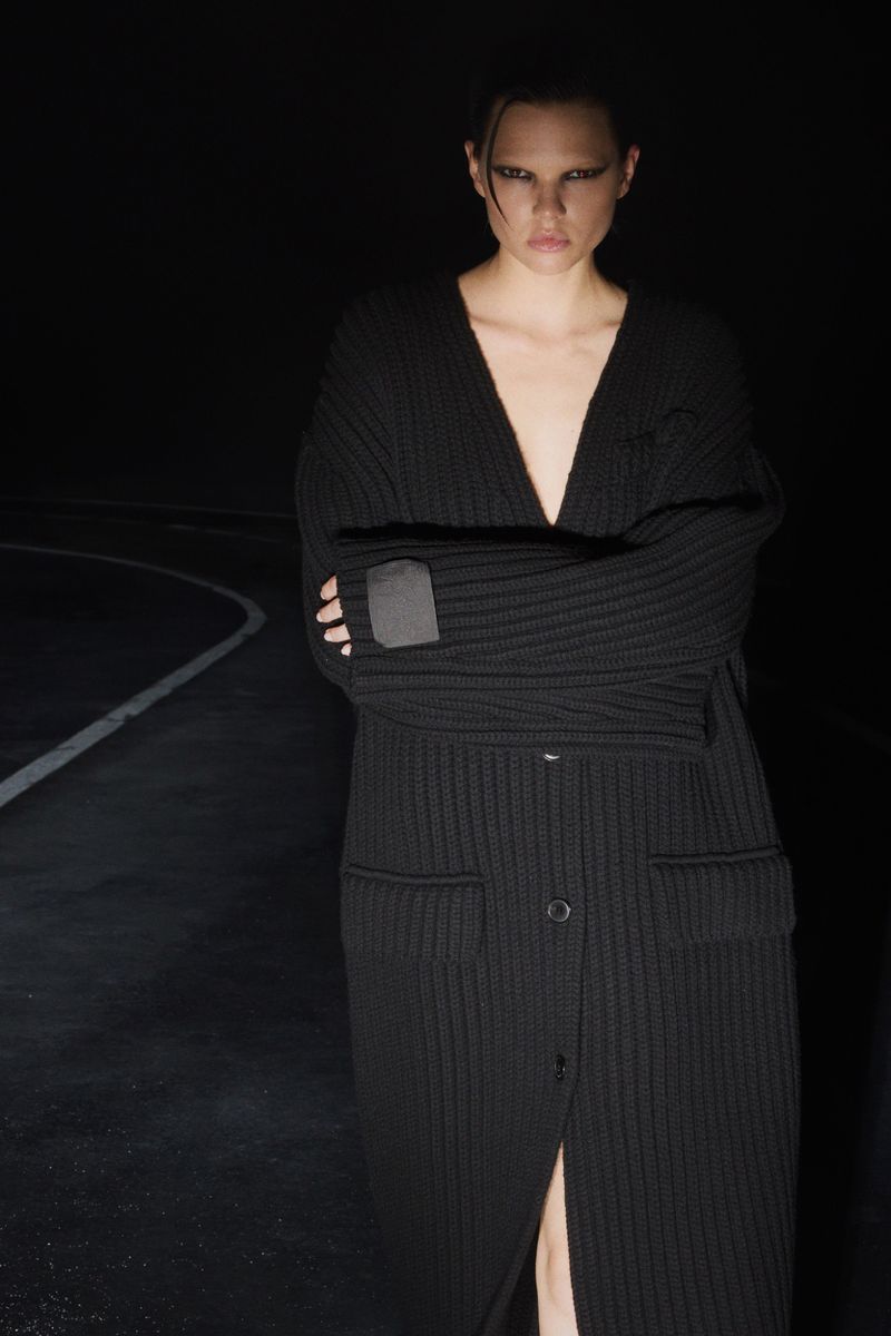 Adult female fashion model posing in long, buttoned, black cardigan in dark indoor setting with graphic eye makeup and slicked-back hair.
