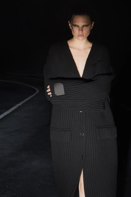 Adult female fashion model posing in long, buttoned, black cardigan in dark indoor setting with graphic eye makeup and slicked-back hair.