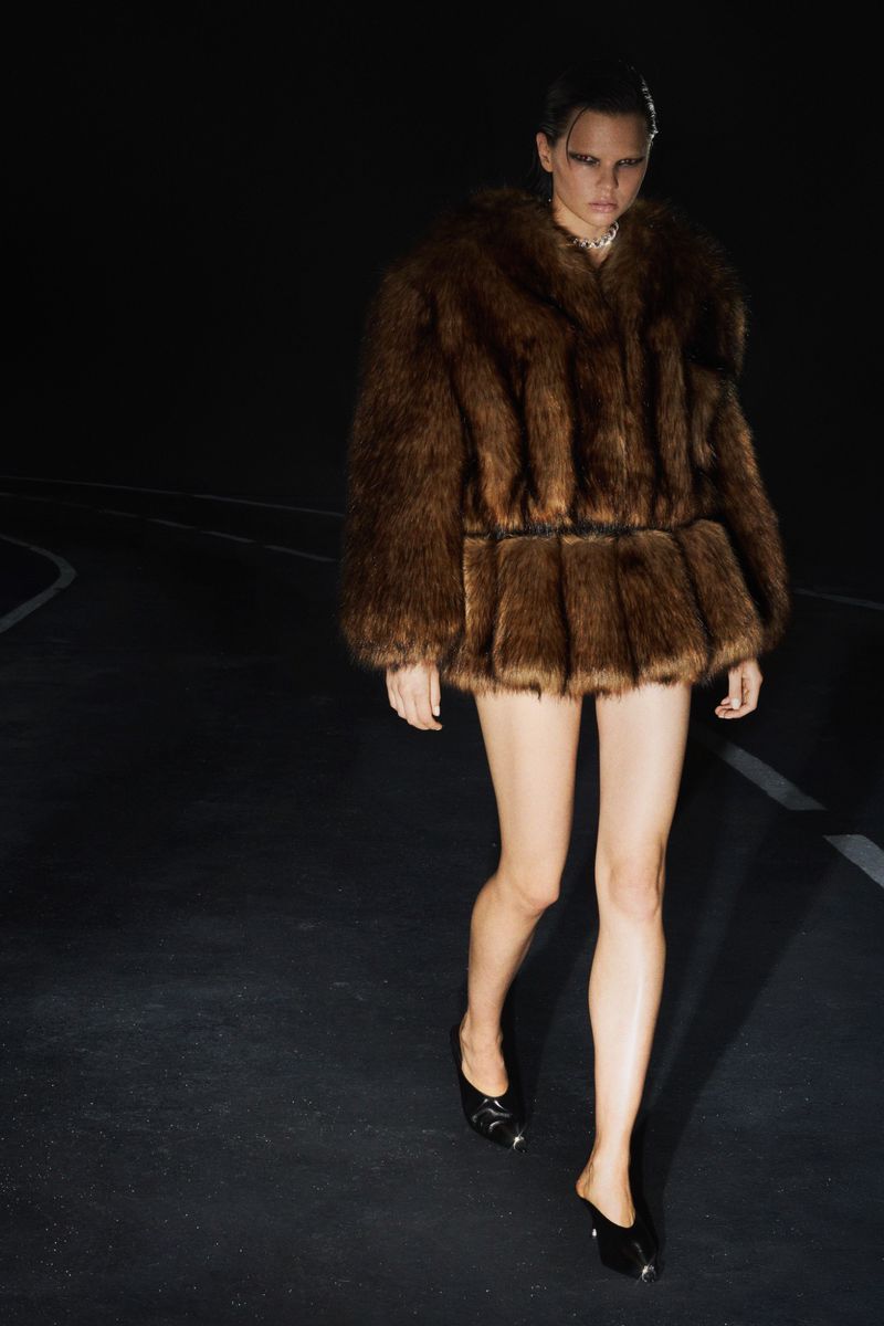 A female model in a brown fur coat and black heels walks on a black asphalt surface with white lines, against a dark backdrop.