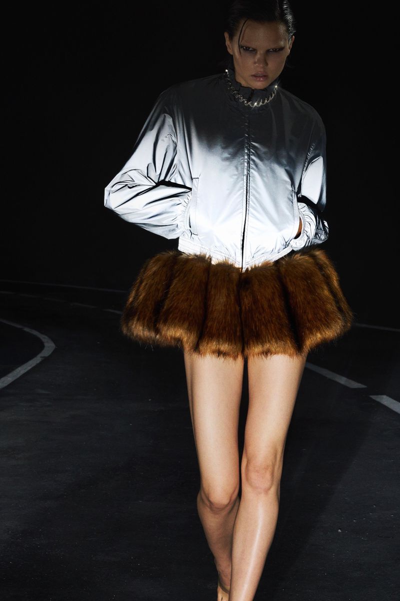A fashion photo features a model in a reflective bomber jacket and brown fur skirt on a dark asphalt runway, captured in a medium shot.