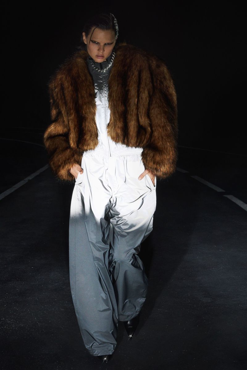 Fashion model poses in an oversized brown faux fur coat and wide-leg grey jumpsuit in a dark studio with directional lighting.