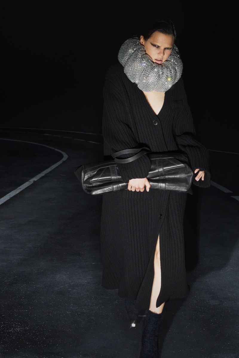 An adult female fashion model wears a silver metal mesh ruff and carries a large black leather bag against a black background.