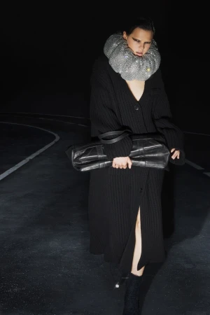 An adult female fashion model wears a silver metal mesh ruff and carries a large black leather bag against a black background.