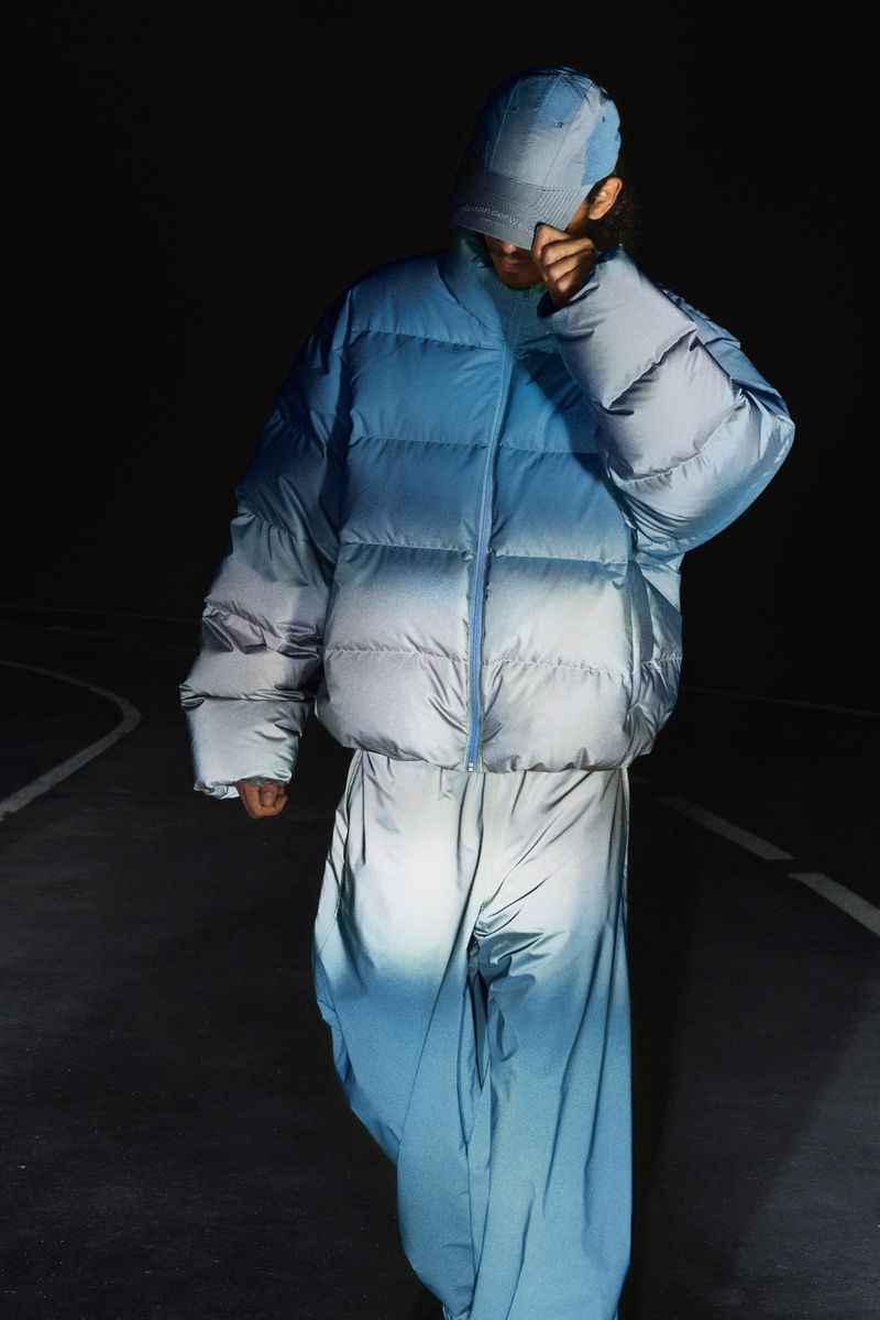 Model wearing a blue ombre puffer jacket, matching trousers, and a cap under low-key night lighting on a dark road.