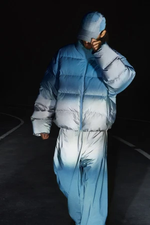 Model wearing a blue ombre puffer jacket, matching trousers, and a cap under low-key night lighting on a dark road.