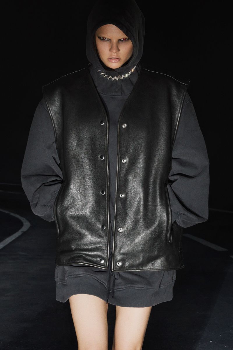 A person with light skin wears a hooded top, a chain necklace, and a black leather vest while standing in front of a dark background.