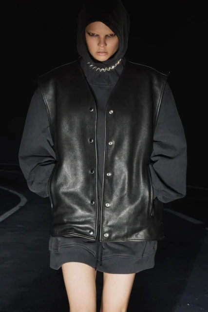 A person with light skin wears a hooded top, a chain necklace, and a black leather vest while standing in front of a dark background.
