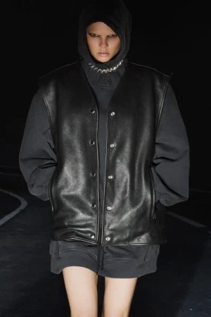 A person with light skin wears a hooded top, a chain necklace, and a black leather vest while standing in front of a dark background.