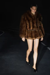Model in brown faux fur jacket and black mules walking on dark asphalt road with lane markings