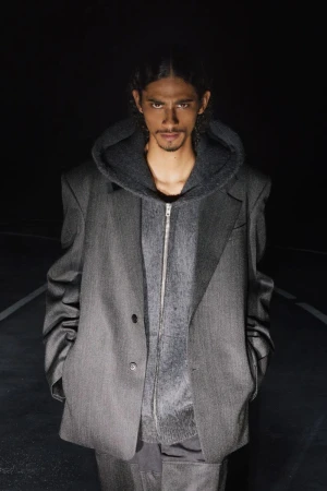 Model wearing an oversized grey tailored blazer layered over a grey zip-up hoodie in a dark outdoor setting.