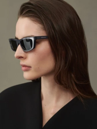 Close-up profile of a woman wearing black angular sunglasses and a dark top against a neutral grey studio background