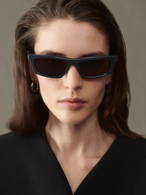 Portrait of a woman with sunglasses wearing a black top, set against a neutral gray background in a studio setting. Fashionable and sleek.