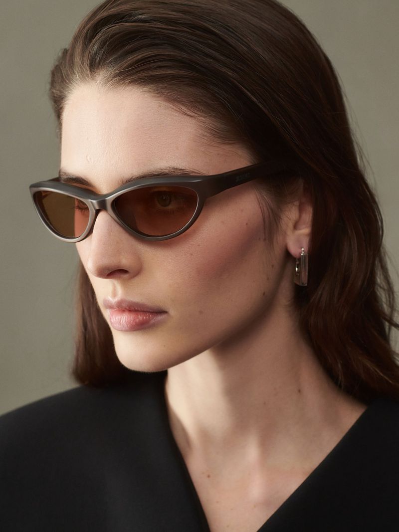 Close-up shot of a brunette woman wearing sunglasses and geometric earrings. The lighting is soft and neutral, highlighting her features.