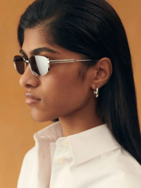 Elegant portrait of a woman with dark hair and white button-down shirt wearing stylish silver sunglasses and hoop earrings on tan backdrop.
