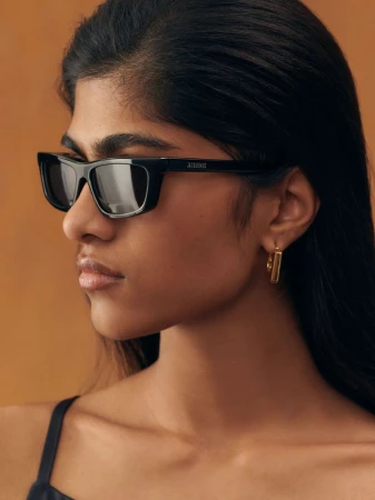 Close-up profile of a model wearing black rectangular sunglasses and gold hoop earrings against a warm brown background