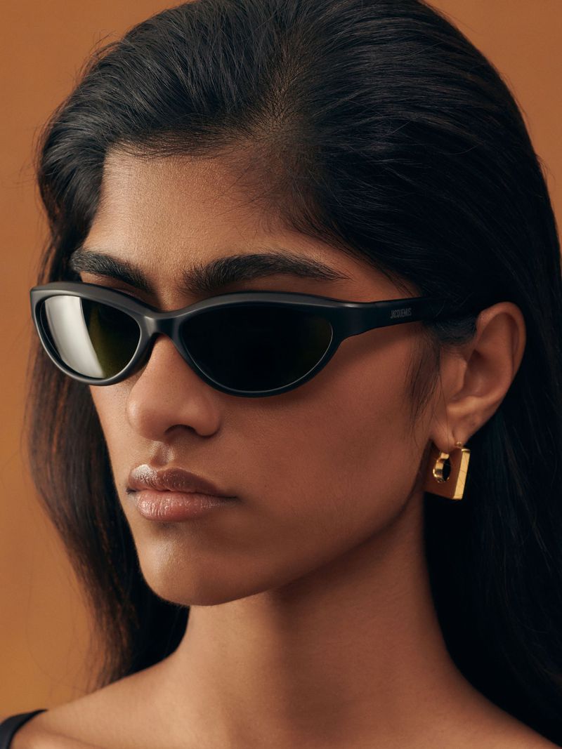 Close-up portrait of a woman wearing black oval sunglasses and a gold geometric earring against a warm brown background.