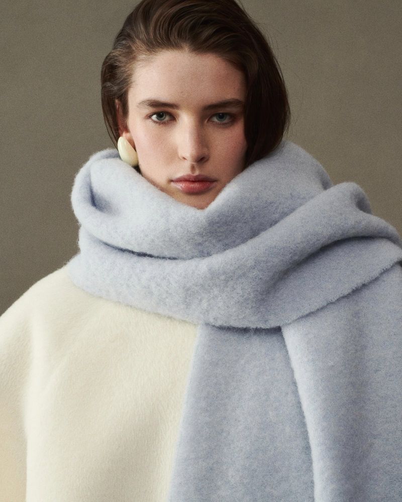 Portrait of an adult woman wearing a large light blue scarf and ivory sweater, posing against a neutral backdrop in a studio setting.