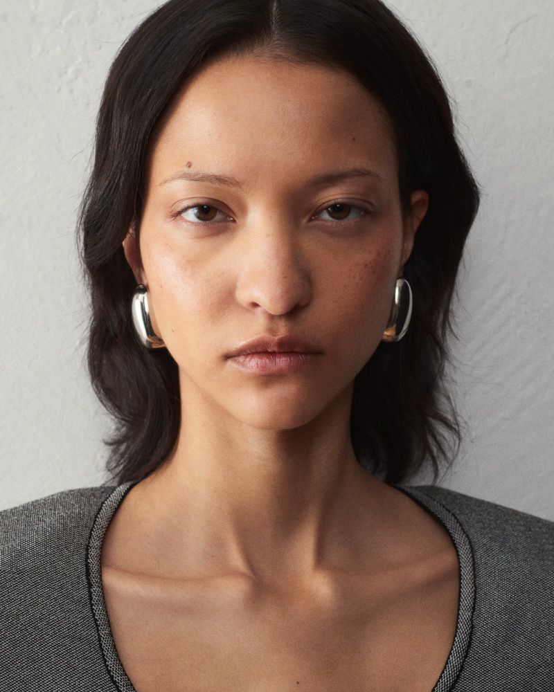 Close-up portrait of a woman wearing chunky silver hoop earrings and a textured grey top against a light wall for Jacquemus.