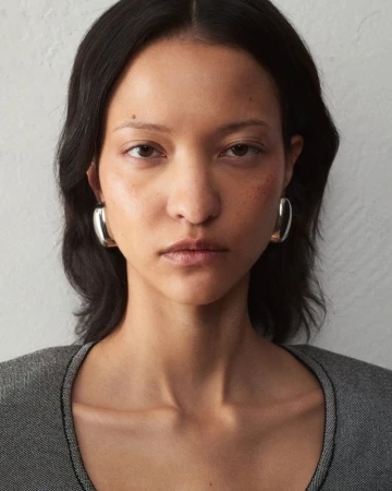 Close-up portrait of a woman wearing chunky silver hoop earrings and a textured grey top against a light wall for Jacquemus.