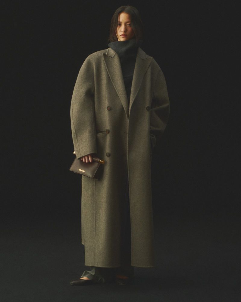 A woman in a wool coat and dark turtleneck poses against a dark backdrop, holding a small dark purse in a studio-lit fashion shot.