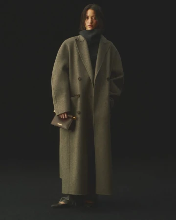 A woman in a wool coat and dark turtleneck poses against a dark backdrop, holding a small dark purse in a studio-lit fashion shot.