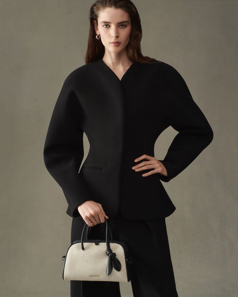 Model in black jacket holding a white and black handbag against a grey background for Jacquemus F/W 25 Lookbook