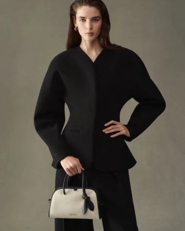 Model in black jacket holding a white and black handbag against a grey background for Jacquemus F/W 25 Lookbook