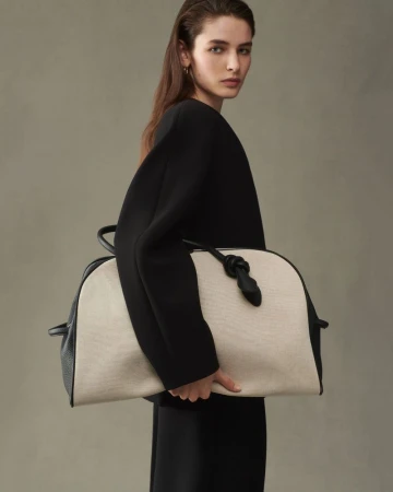 A fashionable adult female model poses in a studio environment holding a neutral-colored, structured handbag in a medium shot.
