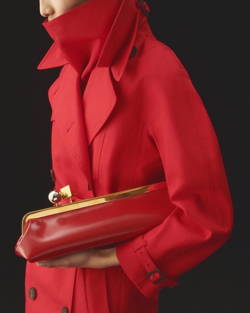 Fashionable adult in a red trench coat holds a red and gold clutch against a dark background in a studio fashion shot.