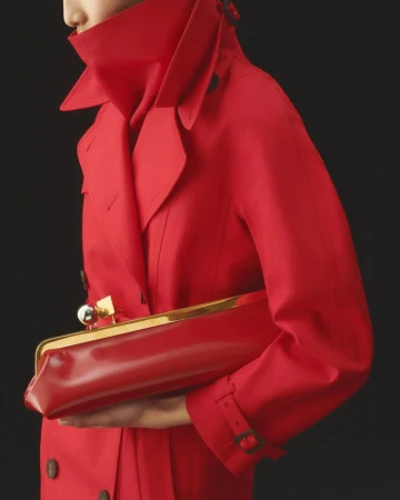 Fashionable adult in a red trench coat holds a red and gold clutch against a dark background in a studio fashion shot.
