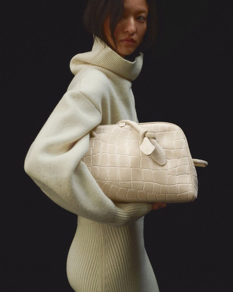 Elegant woman wearing a cream-colored turtleneck sweater dress and holding a matching textured leather handbag against a dark backdrop.