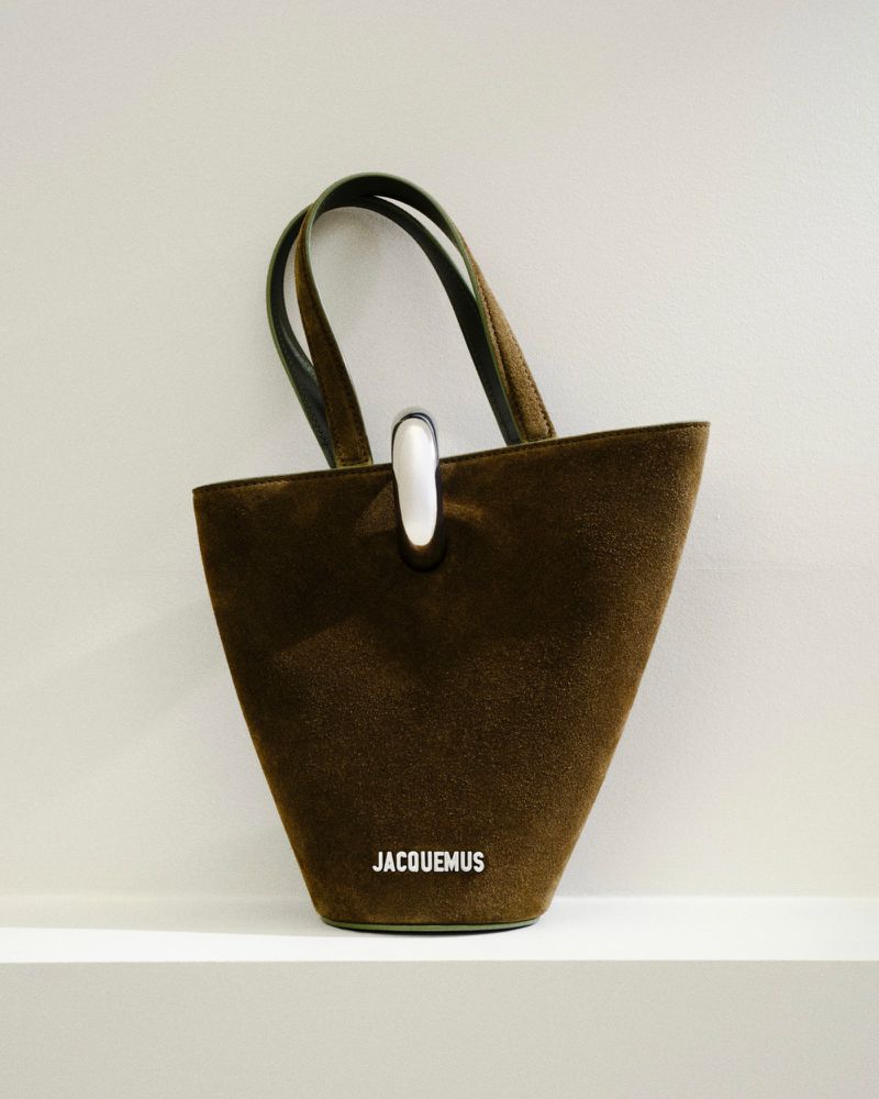 Close-up of a brown suede Jacquemus handbag with a silver handle, resting on a white shelf against a white wall. Fashion.