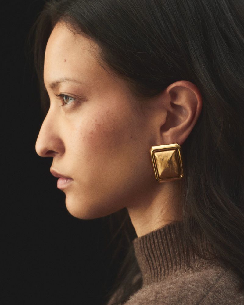 Side profile of a woman wearing a large geometric gold earring and a brown knit turtleneck sweater against a dark black background.