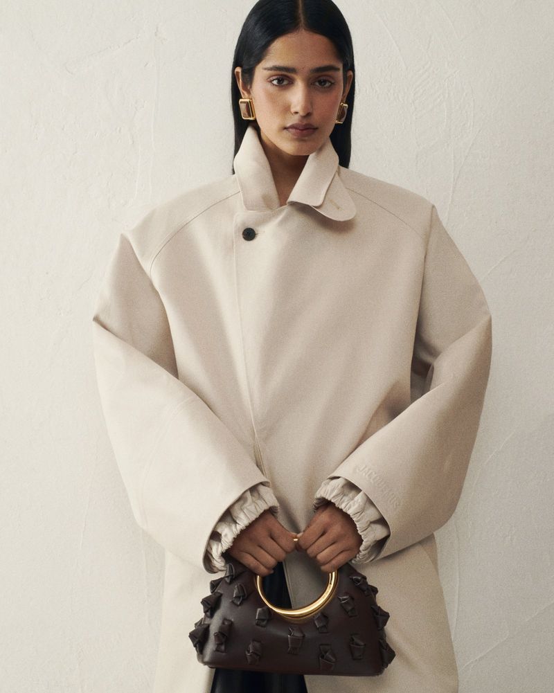 Brunette woman in a tan trench coat holds a structured dark brown leather handbag with a gold circular handle against a cream background.
