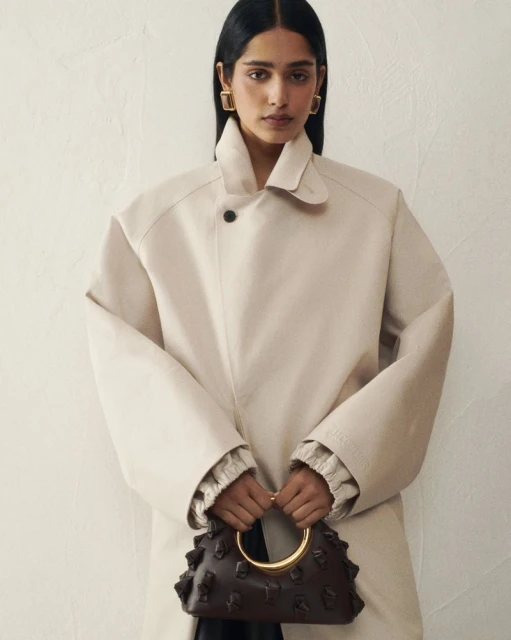 Brunette woman in a tan trench coat holds a structured dark brown leather handbag with a gold circular handle against a cream background.