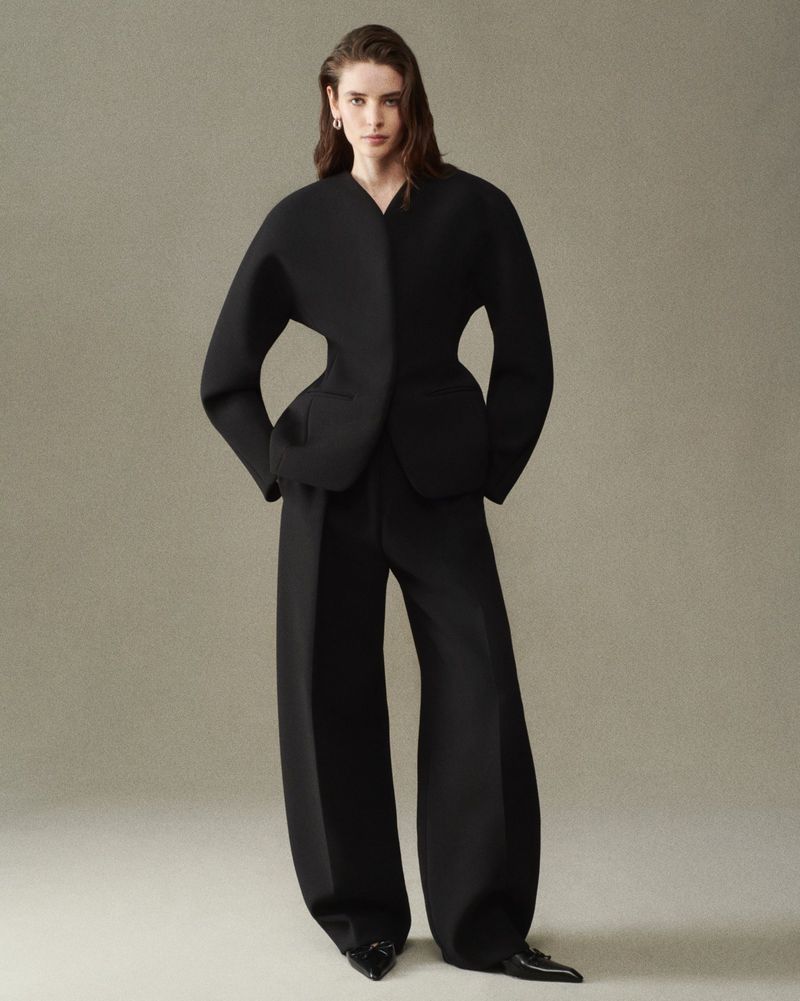 Model wearing a sculptural black suit jacket with wide-leg trousers and pointed shoes against a neutral backdrop.