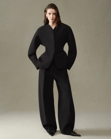 Model wearing a sculptural black suit jacket with wide-leg trousers and pointed shoes against a neutral backdrop.