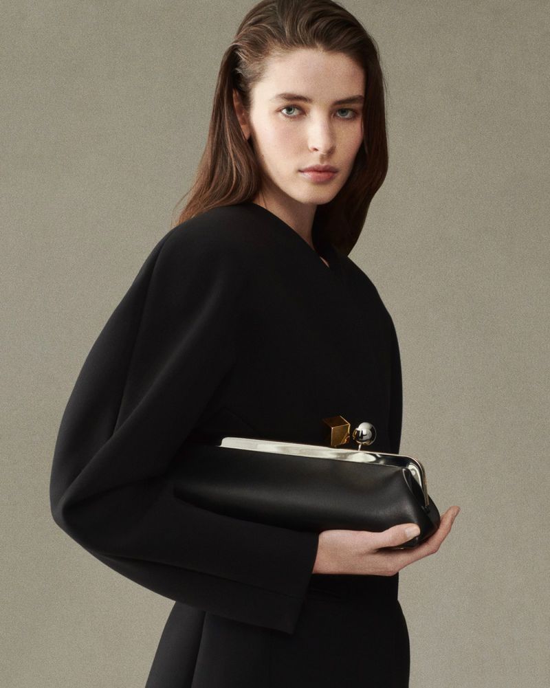 Woman in black dress holds a black leather clutch bag in a studio shot, showcasing fashion and beauty with a neutral backdrop.