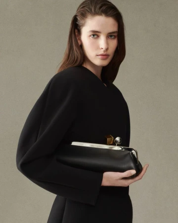 Woman in black dress holds a black leather clutch bag in a studio shot, showcasing fashion and beauty with a neutral backdrop.