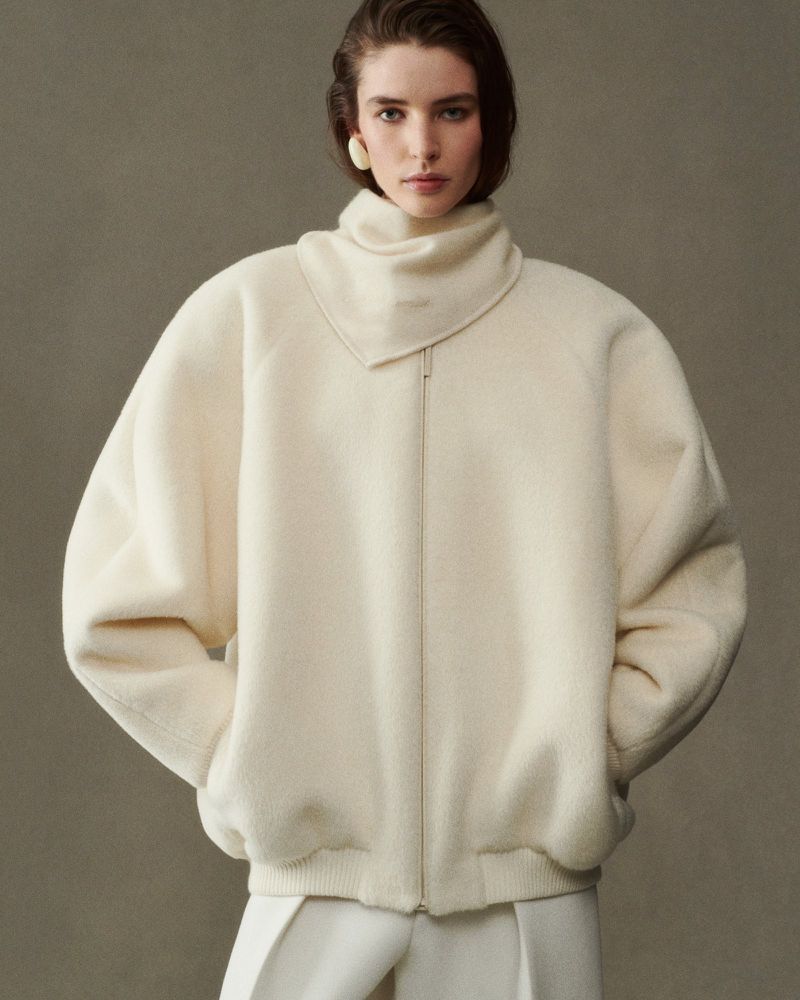 Elegant studio shot of a young woman in a cream-colored sweater with matching trousers, styled with statement earrings, against a neutral backdrop.