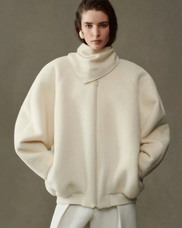 Elegant studio shot of a young woman in a cream-colored sweater with matching trousers, styled with statement earrings, against a neutral backdrop.