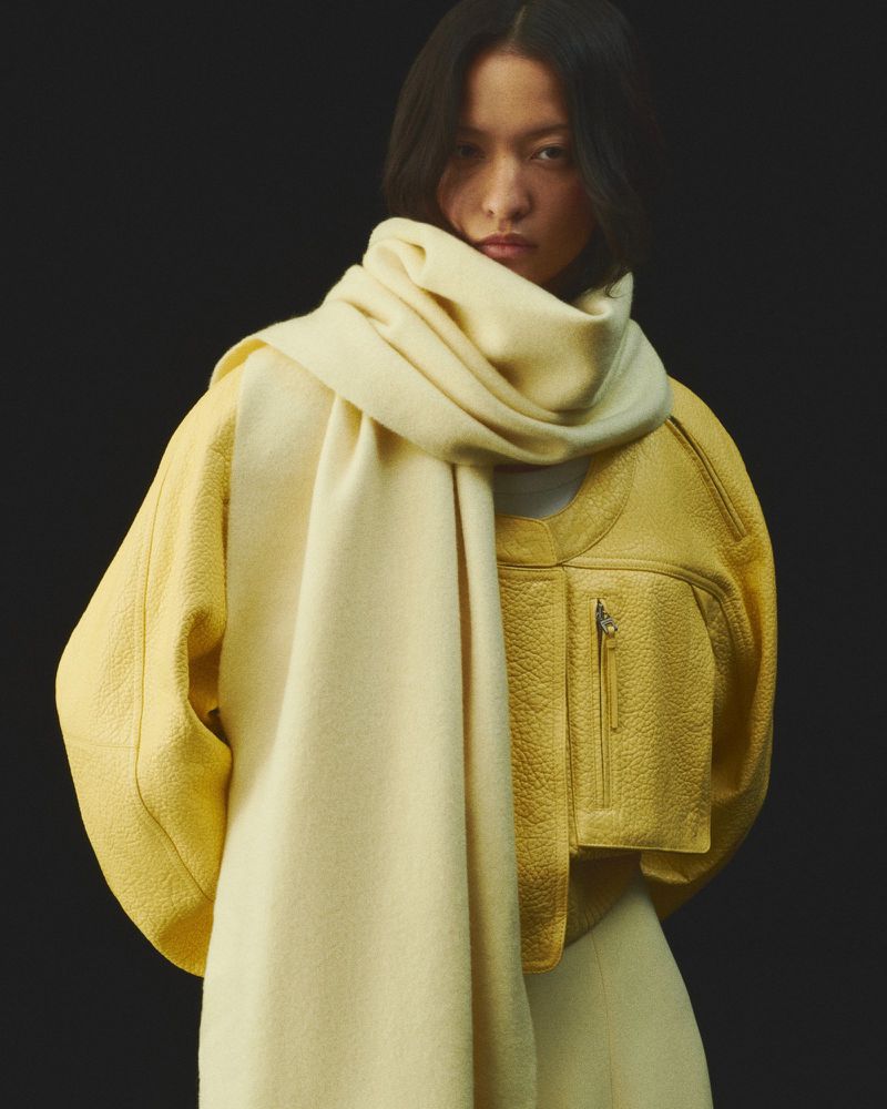 Asian adult woman wearing a yellow jacket and a cream-colored scarf posing in a studio setting with dark background. Fashion shot.