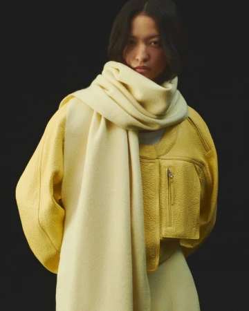 Asian adult woman wearing a yellow jacket and a cream-colored scarf posing in a studio setting with dark background. Fashion shot.