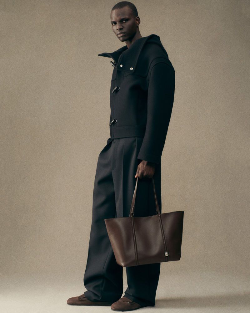 A single adult male stands in a studio wearing a black jacket and pants holding a brown leather tote bag.