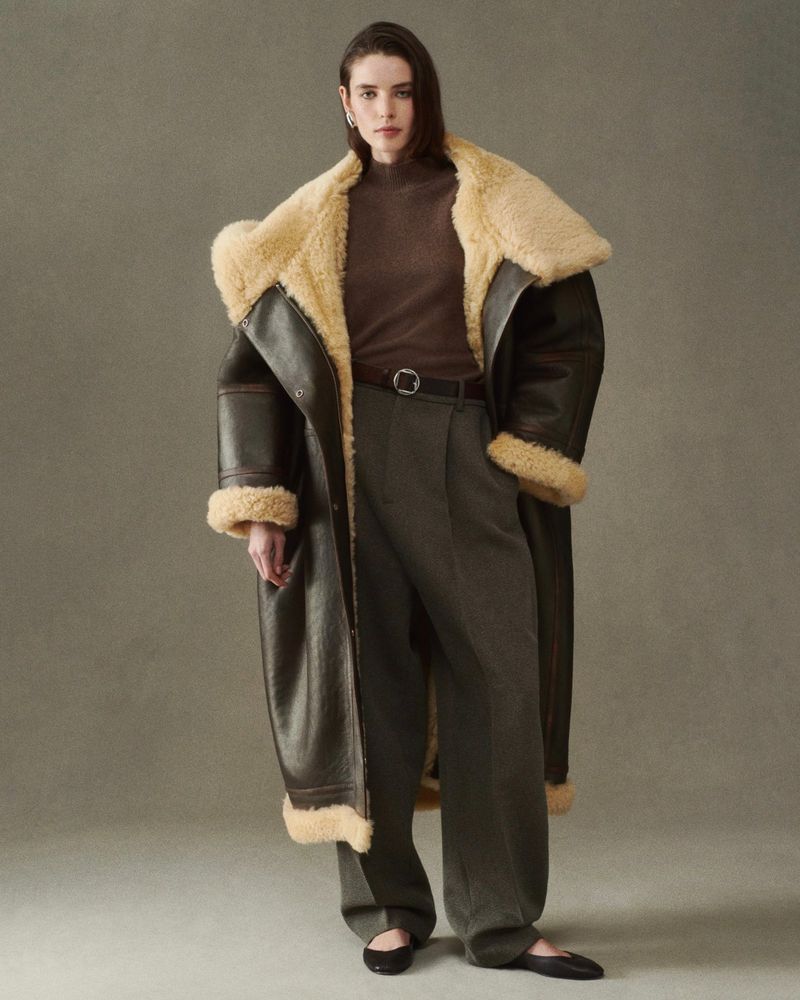 Woman in a brown shearling coat with a turtleneck and wide leg trousers, posing in front of a neutral backdrop.