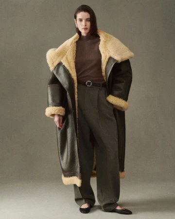 Woman in a brown shearling coat with a turtleneck and wide leg trousers, posing in front of a neutral backdrop.