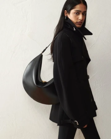 Model wearing a high-collar black jacket carrying a sculptural black leather crescent shoulder bag against a textured wall.